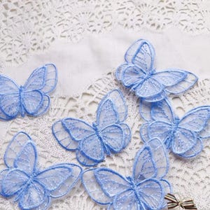 May include: Several light blue butterfly appliques are arranged on a white lace surface. The butterflies have delicate, see-through wings with detailed stitching. The appliques are ideal for crafting and embellishing various items.