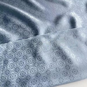 May include: Close-up of a light blue fabric with a floral pattern. The fabric has a subtle sheen and a textured surface. The floral design is repeated across the material, creating a consistent pattern. The edge of the fabric is visible.