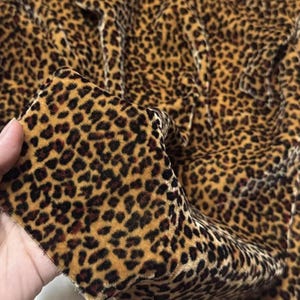May include: A close-up of leopard print fabric. The fabric has a tan base with dark brown spots. The material appears to be velvet or a similar soft fabric. The fabric is held up, displaying its texture and pattern in detail.