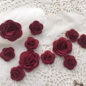 May include: Burgundy fabric rose appliques of varying sizes are displayed on a white lace surface. The image shows several individual roses, ideal for craft projects. The background includes a white feather. These decorative floral embellishments are suitable for crafts and sewing projects.