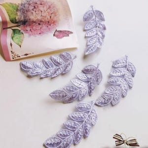 Peut inclure: Plusieurs applications de feuilles brodées de couleur lavande sont disposées sur une surface blanche. Les feuilles présentent des nervures détaillées et sont faites d'un tissu délicat et transparent. Une carte florale avec un papillon est en arrière-plan.