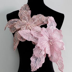 May include: Two floral appliques in shades of pink and rose gold, embellished with sequins and beads. The delicate, layered petals create a three-dimensional effect. These decorative appliques are ideal for embellishing garments or accessories.
