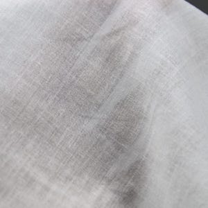 May include: Close-up of a white, semi-transparent fabric with a textured weave. The fabric appears lightweight and slightly creased, with visible diagonal and horizontal lines. The overall tone is a soft, neutral white.