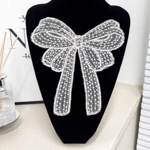 May include: A white, beaded bow applique with a delicate, lacy design. The bow is displayed on a black velvet mannequin, highlighting its intricate details and texture. The bow's design features small, round beads.