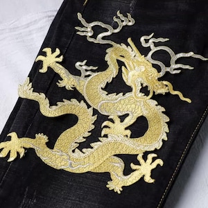 Peut inclure: Gros plan sur un tissu denim noir avec un grand motif de dragon brodé en or. Le dragon est finement détaillé avec du fil d'or et d'argent, créant un effet texturé et accrocheur. Le dragon est centré sur le tissu.
