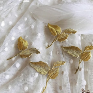 Puede incluir: Cuatro ramitas de hojas doradas bordadas sobre un fondo de tela blanca con lunares. Las hojas tienen costuras detalladas y un brillo metálico. Una pluma blanca está en el fondo. Ideal para decoración o proyectos de manualidades.