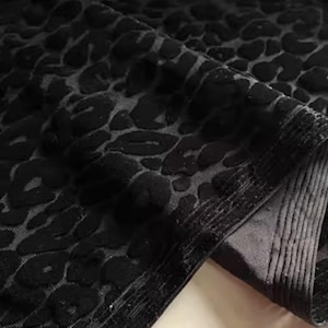 Puede incluir: Primer plano de una tela negra con un estampado de leopardo en relieve. La tela está doblada, revelando un borde texturizado. El diseño presenta formas oscuras e irregulares sobre un fondo ligeramente más claro, creando un contraste.