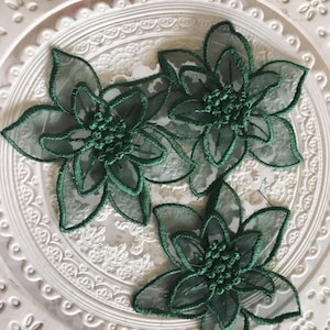 May include: Three dark green floral appliques. Each applique features a sheer, petal-shaped design with a detailed, embroidered outline. The center of each flower is a cluster of textured, dark green material. The appliques are placed on a white, decorative surface.