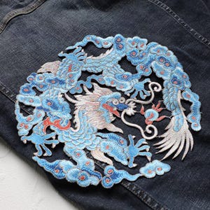 Puede incluir: Parche bordado con un diseño detallado de dragón azul y blanco. El dragón, con detalles en rojo, se encuentra sobre un fondo de mezclilla oscura. El parche, de forma circular, está diseñado para aplicarse en ropa.
