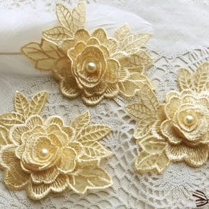 Pode incluir: Três apliques de flores de renda dourada com centros de pérolas e detalhes de folhas. Os enfeites florais são dispostos sobre uma superfície de renda branca, adequados para artesanato ou para adornar roupas.