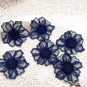 May include: Six navy blue lace flower appliques with a detailed design. The flowers have a layered petal effect and a central, textured core. They are arranged on a white, patterned surface, creating a delicate and elegant aesthetic.
