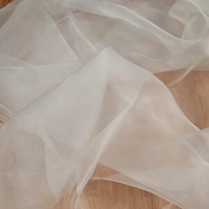 Puede incluir: Primer plano de una tela blanca transparente con una apariencia fluida y drapeada. La tela es translúcida, permitiendo el paso de la luz, y tiene una textura suave y delicada. El fondo es una superficie de madera marrón claro.