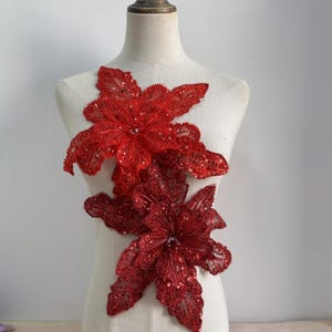 Könnte beinhalten: Zwei rote, paillettenbesetzte Blumenapplikationen. Die Applikationen haben die Form von Weihnachtssternen, mit mehreren Blütenblättern und einer zentralen Verzierung. Der Stoff hat eine zarte, spitzenartige Textur, und die Pailletten verleihen einen Hauch von Glanz. Diese Applikationen eignen sich zum Verzieren von Kleidungsstücken oder Bastelarbeiten.