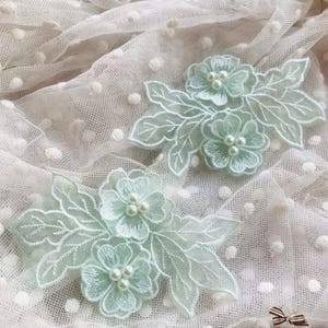 May include: Two pale mint green floral lace appliques featuring embroidered leaves and petals with small white faux pearl centers. The delicate lace motifs are layered over a sheer white fabric with a subtle raised polka dot texture pattern.