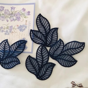 May include: Navy blue leaf-shaped appliques with delicate vein detailing. The appliques are arranged on a white surface, with a floral illustration in the background. These decorative appliques are ideal for embellishing clothing, accessories, or craft projects.