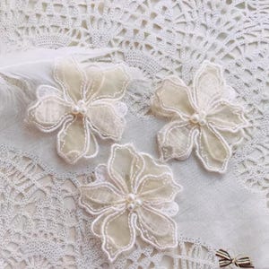 Puede incluir: Tres apliques en forma de flor de color marfil con delicadas costuras blancas y un centro de cuentas de perlas. Los apliques están hechos de una tela transparente y están dispuestos sobre una superficie de encaje blanco, lo que sugiere un tema de artesanía o adorno.