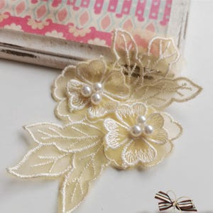 Peut inclure: Applique florale en dentelle ivoire avec trois perles sur chaque fleur. L'applique présente des feuilles et des fleurs délicates, parfaites pour embellir des vêtements ou des créations artisanales. La dentelle est de couleur blanc cassé.