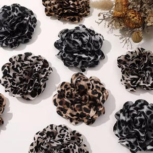 May include: An assortment of fabric flower hair accessories in various shades of brown, black, and white. The flowers have a leopard print pattern. Some flowers are a solid color, while others have a mix of colors.
