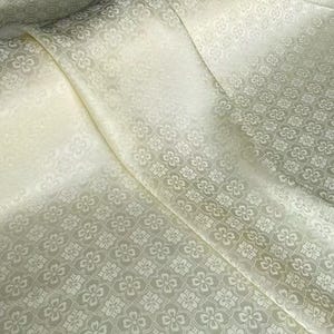 May include: Off-white fabric with a repeating floral pattern. The fabric has a subtle sheen and is draped in soft folds, creating a textured appearance. The floral design is a lighter shade of off-white.
