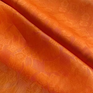 Puede incluir: Primer plano de una vibrante tela naranja con un estampado floral. La tela está doblada, creando sombras y resaltando la textura y el diseño. El patrón presenta motivos florales detallados, agregando profundidad e interés visual al material.