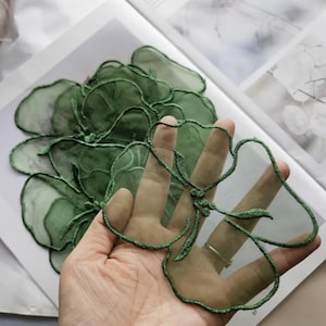 Op de afbeelding: Close-up van groene stoffen bloemenapplicaties met een transparant, gelaagd ontwerp. De applicaties hebben een geborduurde omtrek en worden in een hand gehouden, wat hun ingewikkelde details benadrukt. Deze bloemenversieringen zijn geschikt voor knutsel- en textielprojecten.