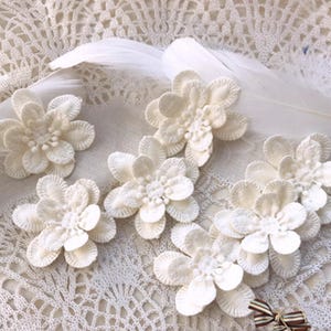May include: A collection of off-white, crocheted flower appliques are displayed on a lace background with a feather. The flowers feature multiple petals and a textured centre. These appliques are suitable for crafting or embellishment.