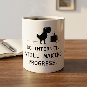 Op de afbeelding: Een witte keramische mok met een zwarte pixelafbeelding van een dinosaurus naast een koffiekopje. Onder de afbeelding staat de tekst: NO INTERNET. STILL MAKING PROGRESS. De mok heeft een cilindrische vorm en een gladde afwerking.