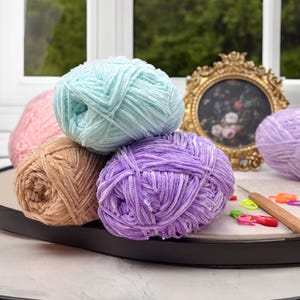 Puede incluir: Una colecci&oacute;n de ovillos de lana coloridos en tonos rosa, turquesa, beige y morado. La lana es suave y esponjosa, perfecta para proyectos de tejido o ganchillo. Un marco dorado y un ganchillo se ven en el fondo.