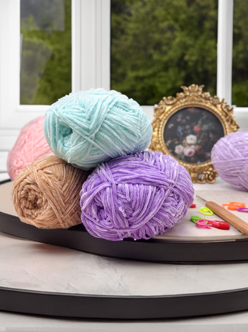 Puede incluir: Una colecci&oacute;n de ovillos de lana coloridos en tonos rosa, turquesa, beige y morado. La lana es suave y esponjosa, perfecta para proyectos de tejido o ganchillo. Un marco dorado y un ganchillo se ven en el fondo.