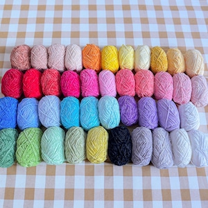 Puede incluir: Una colecci&oacute;n de ovillos de lana coloridos dispuestos en filas. La lana es de varios tonos, incluyendo rosa, naranja, amarillo, azul, verde, morado y blanco. El fondo es un patr&oacute;n a cuadros.