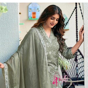 Puede incluir: Un conjunto de salwar kameez verde salvia con un dupatta a juego. El atuendo presenta un estampado floral y ribetes blancos en el dupatta. El conjunto incluye una parte superior con cuello en V y pantalones a juego. La modelo lleva zapatos blancos.