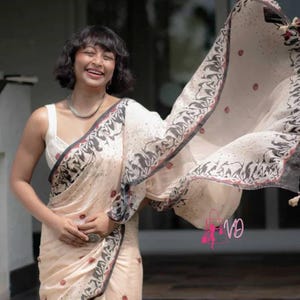 Może przedstawiać: Kremowe sari z dekoracyjnym obramowaniem z czarno-białym wzorem. Sari ma czerwone kwiatowe akcenty i jest narzucone na biały top. Tkanina jest powiewająca, tworząc ruch na zdjęciu. Sari to tradycyjny strój.