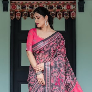 May include: A pink and gray patterned saree with a matching pink blouse. The saree features a floral design and a contrasting border. The blouse has short sleeves and a scoop neckline. The saree is draped over the body.