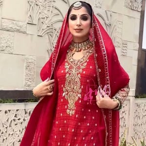 May include: A red embroidered outfit with a matching head covering. The outfit features intricate gold embroidery on the bodice and sleeves. The ensemble includes a necklace and headpiece, creating a formal look. The fabric has a subtle pattern.