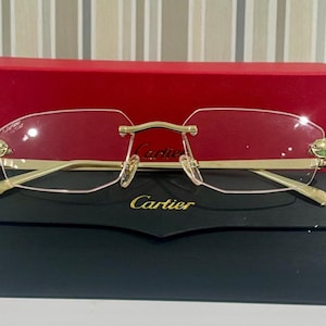 May include: Rimless spectacles with gold-tone frames and a black case. The spectacles have a rectangular shape and are set against a red box. The brand name "Cartier" is visible on the case and the spectacles.
