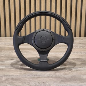 May include: Black steering wheel with a circular center and three spokes. The center features a logo and the word "momo". The wheel has a textured grip and is set against a wooden background.