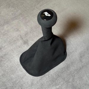 Könnte beinhalten: Schwarzer Autoschaltknauf mit schwarzer Manschette. Der Knauf ist rund und trägt die weißen Buchstaben "RIF" auf einer glänzend schwarzen Oberfläche. Die Manschette besteht aus weichem, schwarzem Stoff.