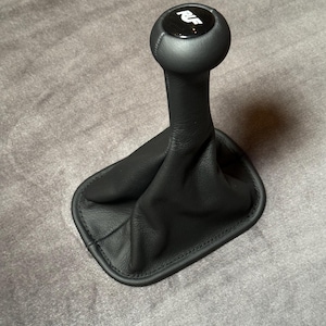 Könnte beinhalten: Schwarzer Lederschaltknauf mit einem schwarz-silbernen Emblem oben. Die Schalthebelmanschette ist ebenfalls schwarz und an den Rändern genäht. Der Schalthebel ist für ein Fahrzeug konzipiert.