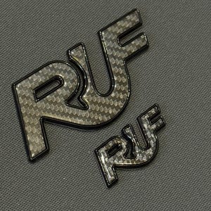May include: Two black and silver RUF emblems, one large and one small, with a carbon fiber weave pattern. The letters R, U, and F are visible. The emblems have a glossy finish and are set against a textured gray background.