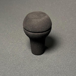 Puede incluir: Un pomo de cambio negro y texturizado con una parte superior redondeada y una base cilíndrica. El pomo está hecho de un material mate y está diseñado para el cambio de marchas de un vehículo.