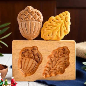 Könnte beinhalten: Eine Holzform mit Eichel- und Blattmotiven, zusammen mit Backwaren in den gleichen Formen. Die Form ist aus hellem Holz gefertigt. Eine kleine Weihnachtsmannfigur und eine Schüssel mit braunem Pulver befinden sich im Vordergrund.