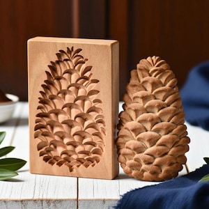 May include: A wooden pine cone mold and a baked pine cone-shaped pastry sit on a white surface. The mold is rectangular with a carved pine cone design. The pastry is golden brown and detailed.