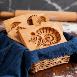 May include: A collection of wooden cookie molds with various carved designs, including a cat and floral patterns, are displayed in a wicker basket with a blue cloth lining. A rolling pin rests in the background, suggesting baking.