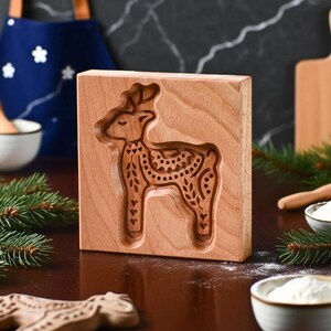 May include: A wooden cookie mold with a carved reindeer design. The rectangular mold is made of light brown wood and features a detailed outline of a reindeer with decorative patterns. The mold is likely used for making Christmas cookies.