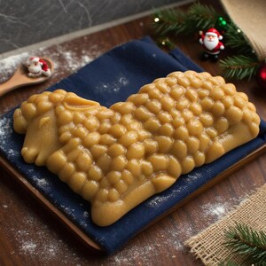 May include: A golden-brown, sheep-shaped pastry sits on a navy blue napkin atop a wooden tray. The pastry has a textured surface and is surrounded by Christmas decorations, including a Santa figurine and greenery.