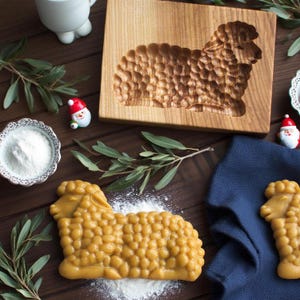 May include: A wooden sheep-shaped cookie mold with a carved design, alongside a baked cookie in the same shape. The image also includes a bowl of white powder, sprigs of greenery, and small Santa Claus figurines, suggesting a holiday baking theme.