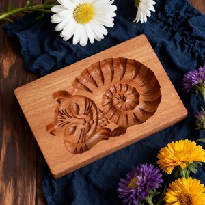 May include: A wooden mold for making cookies or other baked goods. The mold is rectangular and features a carved design of a cat curled up next to a spiral. The wood has a natural, light brown color.