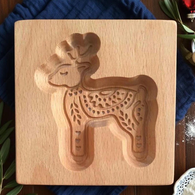 May include: A wooden cookie mold featuring a detailed deer design. The deer is carved into the wood, showcasing intricate patterns on its body and antlers. The mold is square and made of light-colored wood.