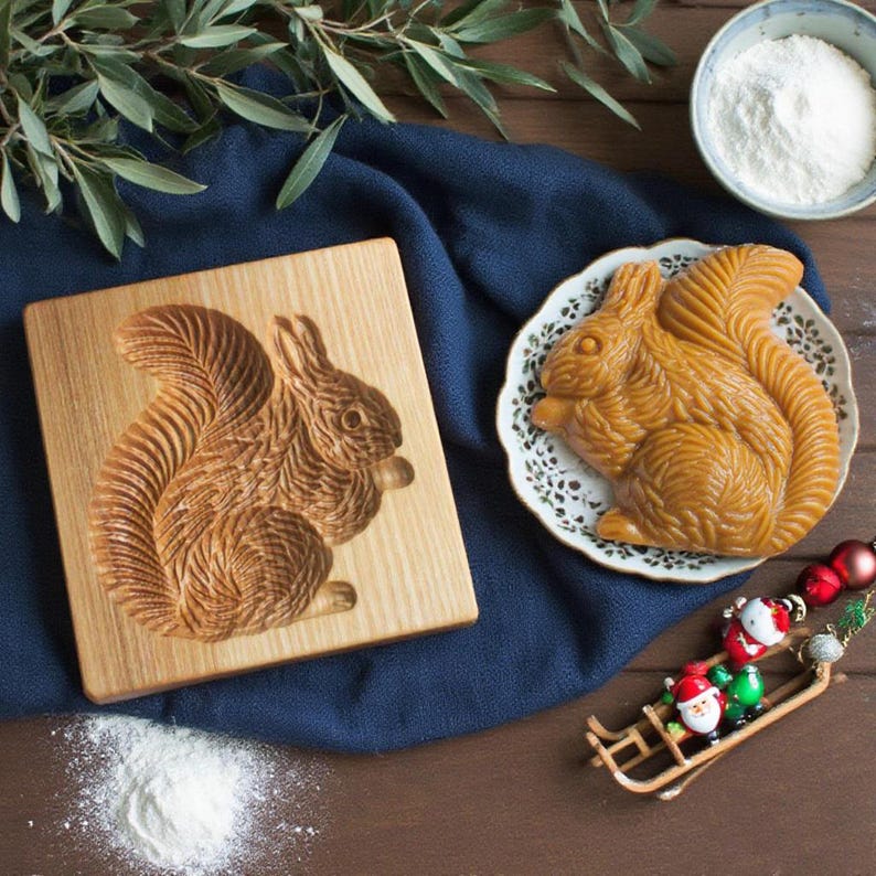 May include: A wooden squirrel-shaped mold and a baked squirrel-shaped pastry on a decorative plate. The mold is carved with intricate details. The pastry is golden brown. A small bowl of white powder and Christmas decorations are also present.