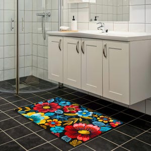 May include: A rectangular bath mat with a floral design. The mat features large red, yellow, and blue flowers with green leaves on a black background. The mat is placed on a black tiled floor in a bathroom setting.